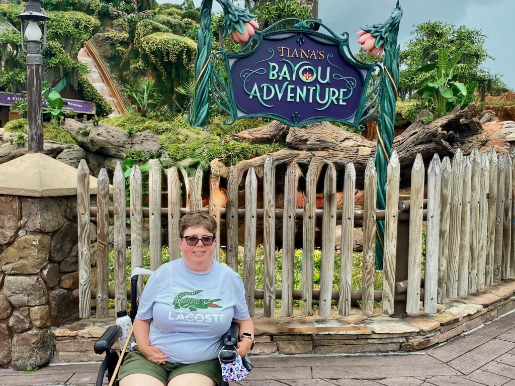 Tiana’s Bayou Adventure Accessibility Review in Walt Disney World Tiana_1 Author in wheelchair with Tiana's Bayou Adventure sign and glimpse of attraction behind her. Author in wheelchair with Tiana's Bayou Adventure sign and glimpse of attraction behind her.